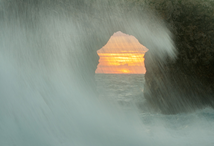 A dramatic sunset photographed through a natural sea arch, with waves crashing in the foreground